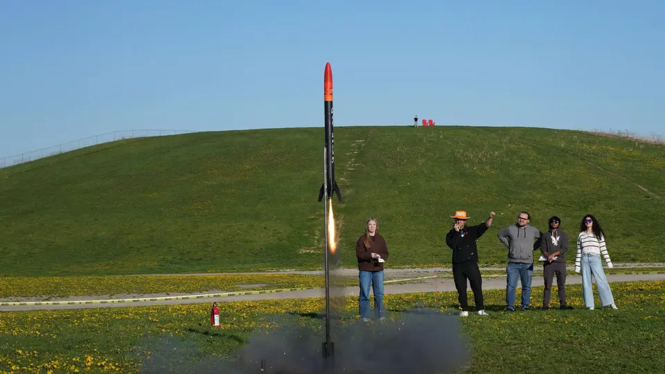 Black rocket lifting off beside students on a grassy field