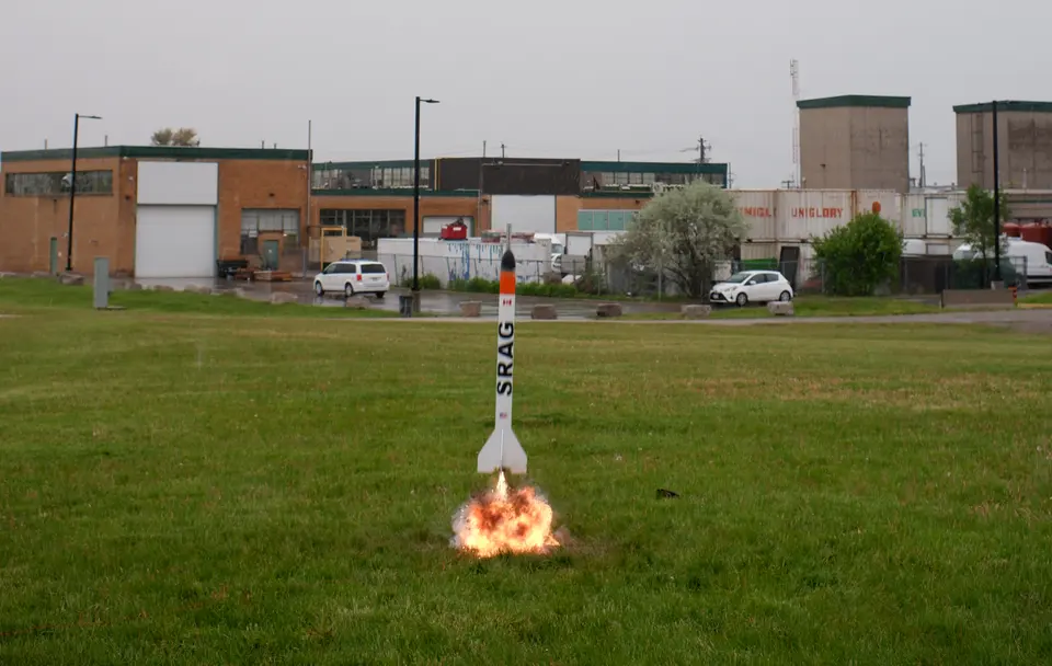 Rocket lifting off a grassy field in front of campus buildings